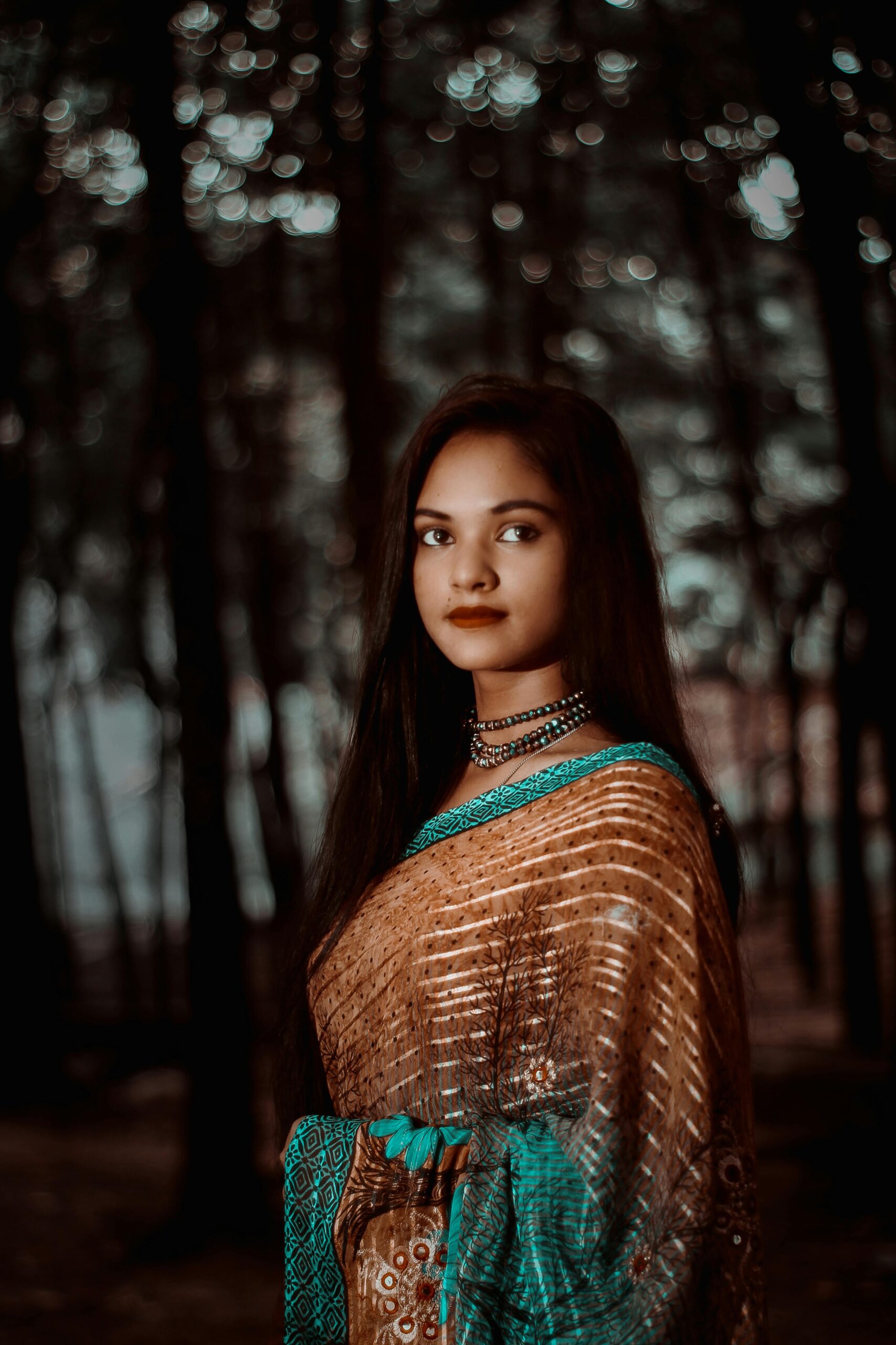 Beautiful woman in traditional saree posing in a serene forest setting, showcasing cultural elegance.